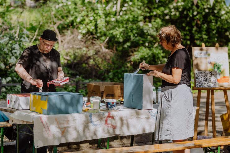 Upcycling Event Zwei Personen bemalen Holzboxen an einem Tisch im Freien, umgeben von Grün und Kunstmaterialien.