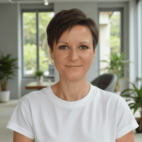 Portrait einer Frau mit kurzem Haar, die in einem modernen Raum mit Pflanzen im Hintergrund lächelt.