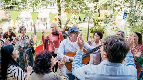 Gruppentanz mit fröhlichen Menschen im Freien, einer spielt Gitarre, umgeben von bunten Dekorationen.