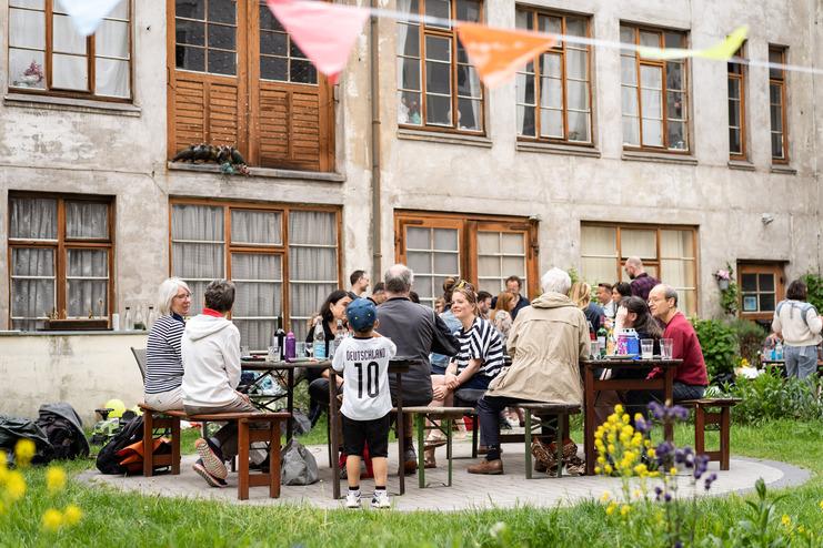 Nachbarschaftsfest Tag der Nachbarn Gruppencollage von Menschen, die an einem Tisch in einem Innenhof sitzen, umgeben von grüner Wiese und bunten Flaggen.