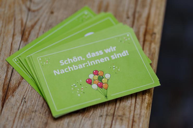 Tag der Nachbarn Grüne Karten mit der Aufschrift „Schön, dass wir Nachbar:innen sind!“ und bunten Luftballons auf einem Holztisch.